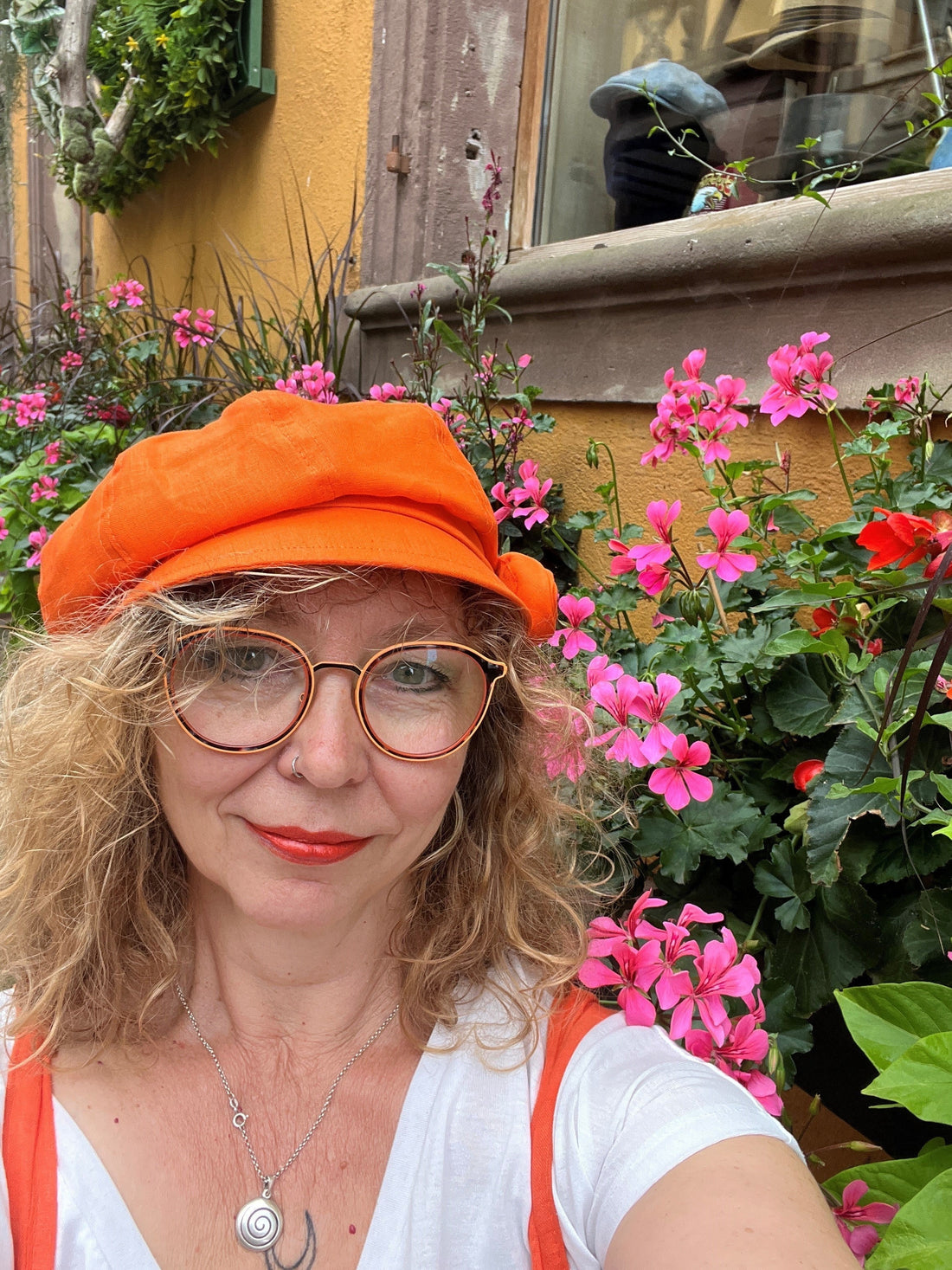 Monika, die Shopinhaberin von Simsalagrün, vor einem Hintergrund mit Geranien, mit orangefarbener Sommermütze