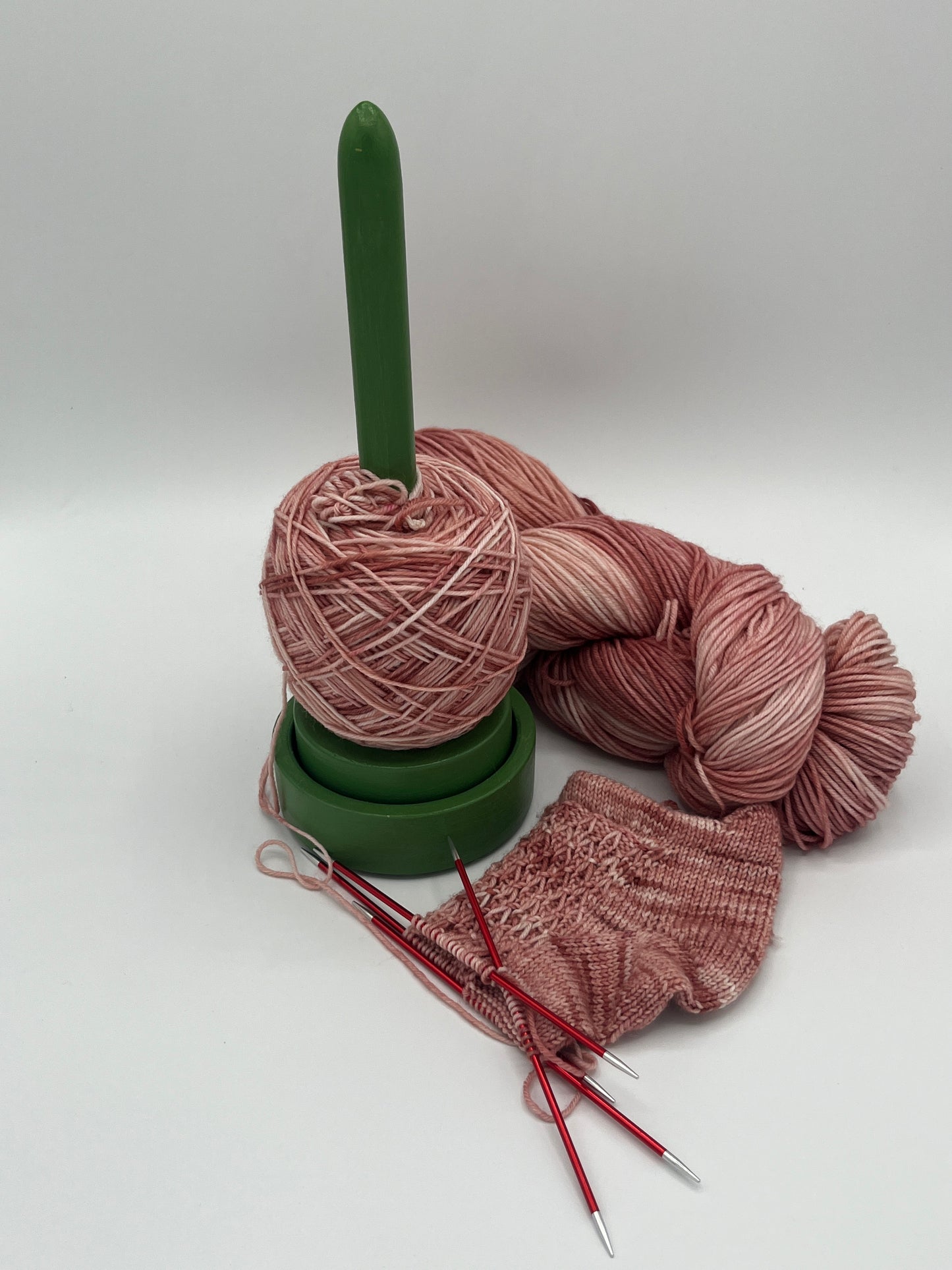 Eine begonnene Sneakersocke Luftikus in der Farbe Rosenprinzessin, also Altrosatöne, mit einem gewickelten Knäuel Wolle auf einem grünen Wollabwickler